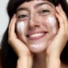 freya-wilson-10 A smiling brunette woman with freckles applies white skincare cream to her face in a studio setting.