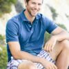 A handsome man with a warm smile wears a blue polo shirt in a bright outdoor setting.