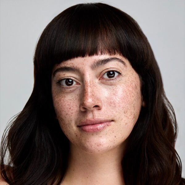 freya-wilson-1 A natural portrait of a young woman with dark hair, bangs, and prominent freckles against a backdrop.