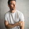 A man with grey-flecked hair in a white t-shirt looks away against a neutral studio backdrop.