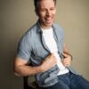 A friendly middle-aged man with a genuine laugh sitting on a chair against a neutral studio backdrop.