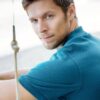 A handsome man with stubble in a teal polo shirt looks back over his shoulder outdoors.