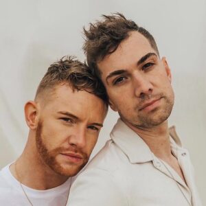 Two men posing closely together in a neutral-toned outdoor field with a large white fabric backdrop.