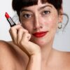 freya-wilson-4 A freckled brunette model wearing vibrant red lipstick poses confidently while holding a lipstick tube near her face.