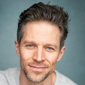 A friendly middle-aged male model with salt and pepper hair smiling warmly against a blue backdrop.