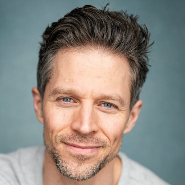 A friendly middle-aged male model with salt and pepper hair smiling warmly against a blue backdrop.