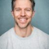 A friendly middle-aged man with a genuine smile poses against a neutral blue studio backdrop for headshots.