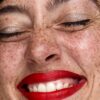 freya-wilson-11 A close up beauty shot of a freckled woman with bright red lipstick laughing with closed eyes.