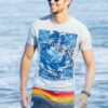 A smiling man wearing sunglasses and a patterned t-shirt walks along the beach in bright sunlight.