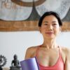 A calm, smiling middle-aged woman holds a purple yoga mat in a bright and peaceful home interior.