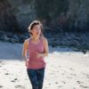 A fit woman in a pink tank top jogs along a sandy beach on a sunny day.