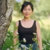 A friendly woman with a warm expression standing outdoors next to a tree in a sunny park.